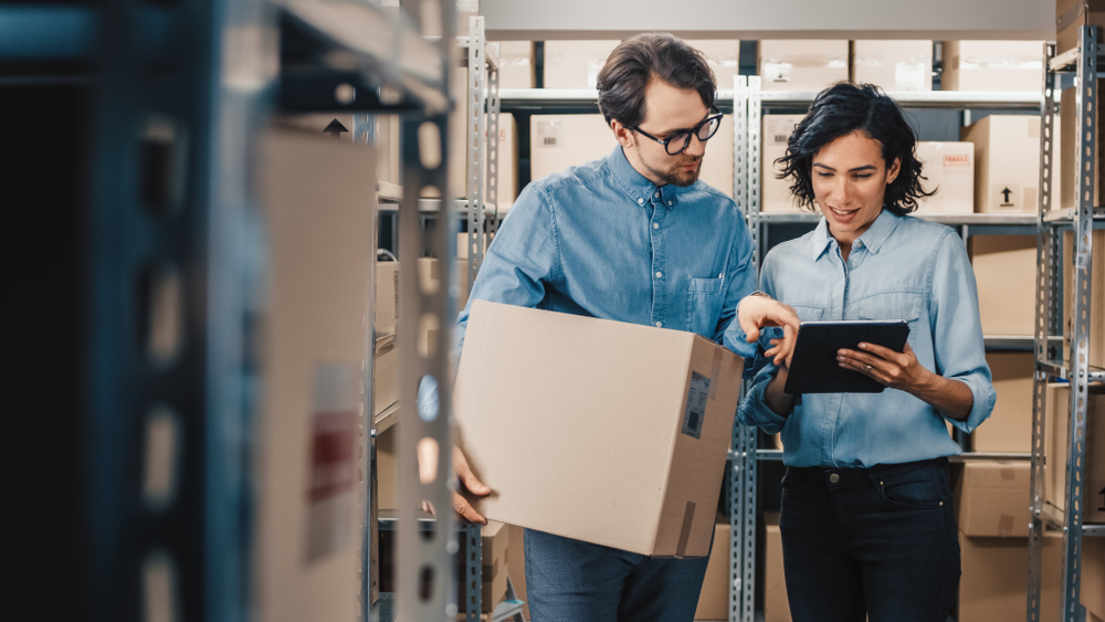 Two employees in a warehouse discussing logistics while holding a box and using a tablet